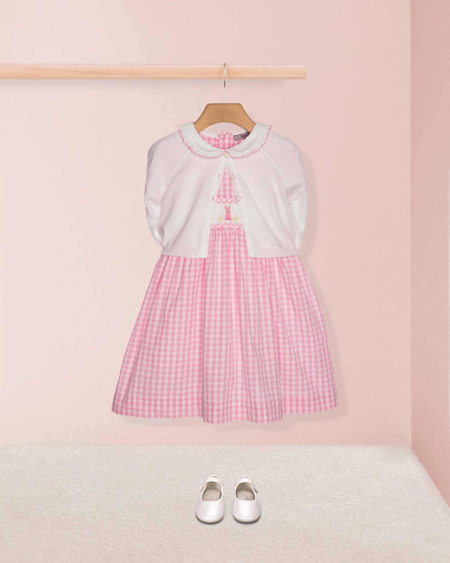 Pink checkered dress with white sweater on a hanger against a pink wall.