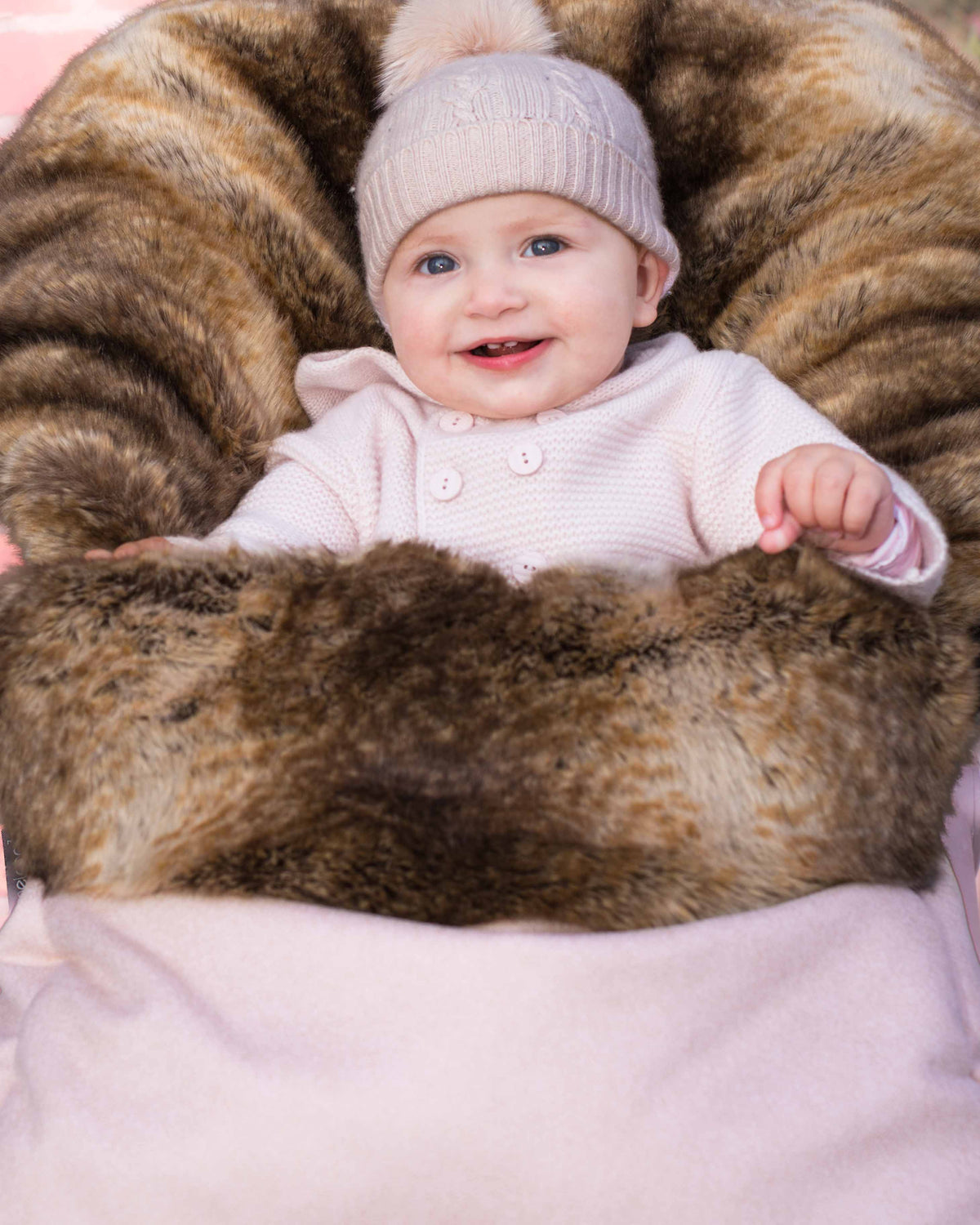 A smiling baby wearing a light pink knit hat with a pom-pom and a pale pink outfit, lying on a brown and black faux fur lounger cushioned by a soft pink blanket.