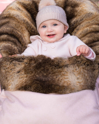A smiling baby wearing a light pink knit hat with a pom-pom and a pale pink outfit, lying on a brown and black faux fur lounger cushioned by a soft pink blanket.