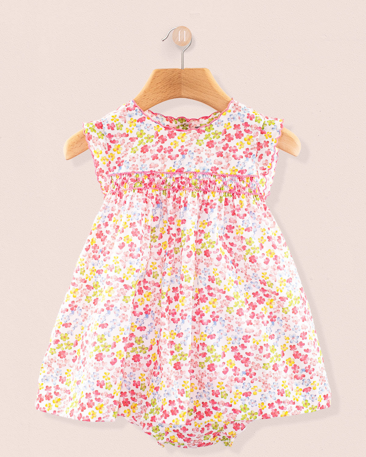 Baby Amelie sleeveless floral dress with smocking and mini collar hanging on a wooden hanger against a neutral wall