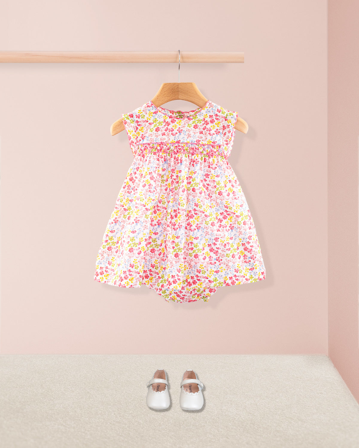 Baby Amelie floral dress shown with matching bloomers and white Mary Jane shoes displayed under a wooden rail