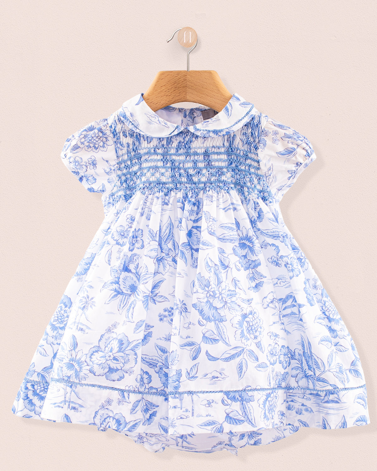 Front view of the Baby Emmy Liberty Delft Blue Dress Set hanging on a wooden hanger. The dress is made from Liberty of London cotton with a soft white background and intricate blue floral print, featuring a fully smocked bodice, puff sleeves, and lace trim at the collar, sleeves, and hem.
