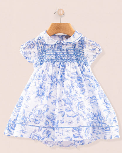 Front view of the Baby Emmy Liberty Delft Blue Dress Set hanging on a wooden hanger. The dress is made from Liberty of London cotton with a soft white background and intricate blue floral print, featuring a fully smocked bodice, puff sleeves, and lace trim at the collar, sleeves, and hem.