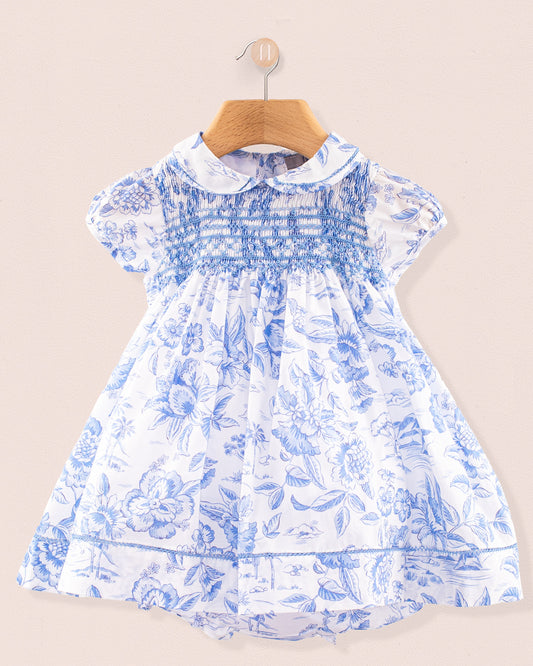 Front view of the Baby Emmy Liberty Delft Blue Dress Set hanging on a wooden hanger. The dress is made from Liberty of London cotton with a soft white background and intricate blue floral print, featuring a fully smocked bodice, puff sleeves, and lace trim at the collar, sleeves, and hem.
