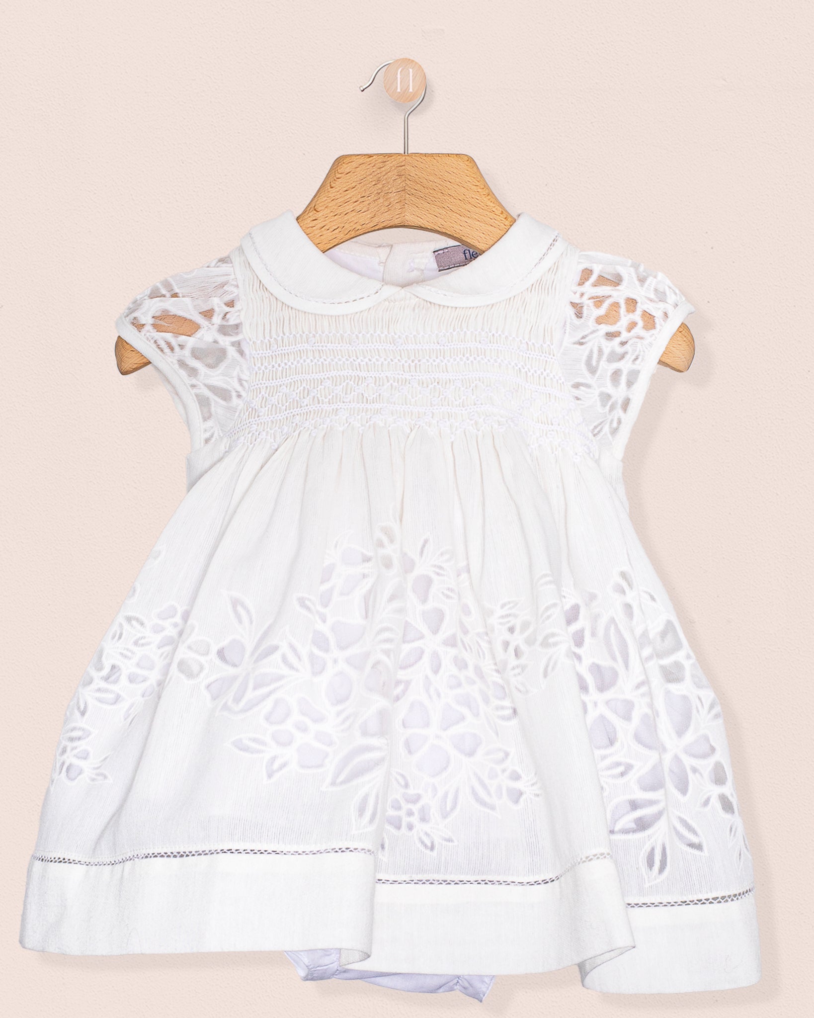 Front view of the Baby Emmy Doppia White Smock Dress Set hanging on a wooden hanger, showcasing floral laser-cut detailing and hand smocking