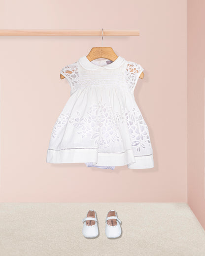 Styled shot of the Baby Emmy Doppia White Smock Dress Set with baby shoes, set against a soft peach background.
