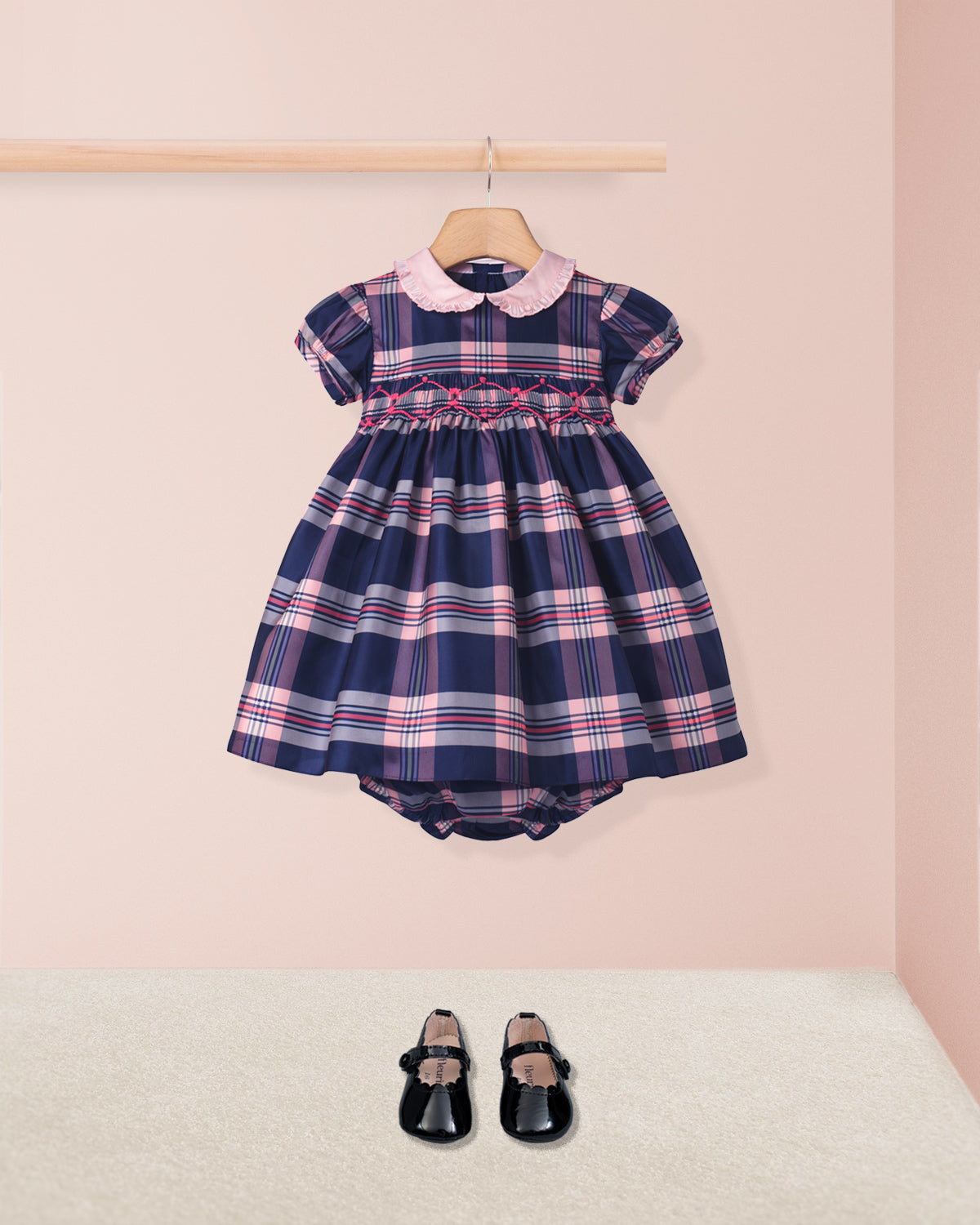 Navy plaid dress with pink accents on a hanger against a light pink wall, with black shoes on a white surface.