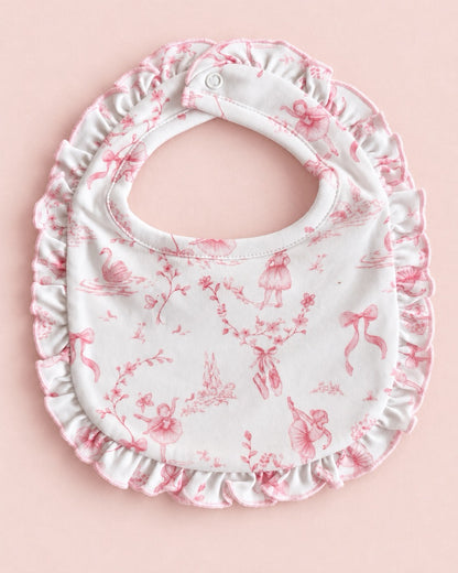 A white fabric baby bib with a pink toile de Jouy pattern featuring ballerinas, bows, and floral motifs, finished with a ruffled edge and snap closure, shown on a solid pale pink background.