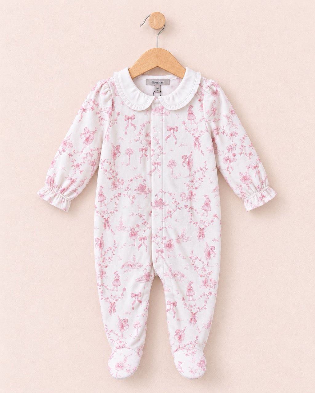 A baby footed one-piece sleeper in white fabric with a pink floral toile pattern, featuring a white Peter Pan collar and long ruffled sleeves, hanging on a wooden hanger against a solid pale pink background.