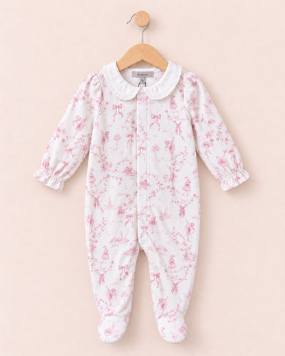 A baby footed one-piece sleeper in white fabric with a pink floral toile pattern, featuring a white Peter Pan collar and long ruffled sleeves, hanging on a wooden hanger against a solid pale pink background.