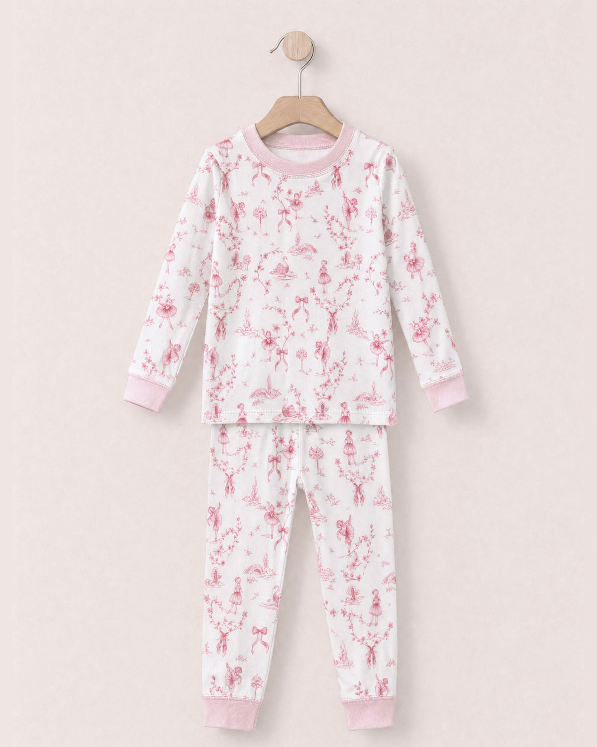 white pajamas with reddish pink toille, bows and ballerinas design, with pink collars and cuffs, on a hanger against a peach neutral background.