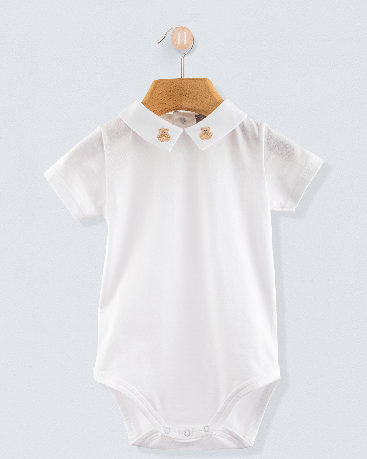 Front view of the Andreas Baby Onesie, a short-sleeve white cotton jersey bodysuit with a pointed collar featuring embroidered beige teddy bears.