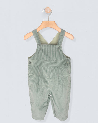Green velvet corduroy overalls hanging on a wooden hanger against a light gray background