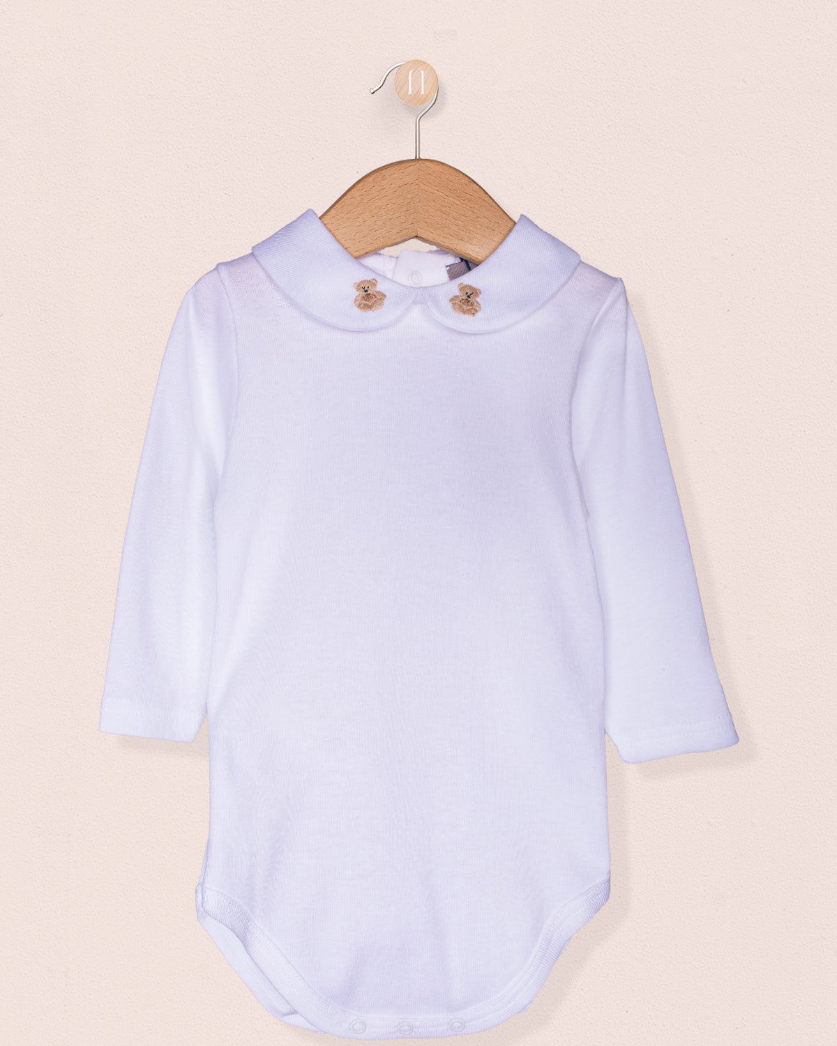 White long-sleeve Italian cotton baby onesie with a beige scalloped-round collar featuring two hand-embroidered beige teddy bears, hanging on a wooden hanger against a soft blush background.
