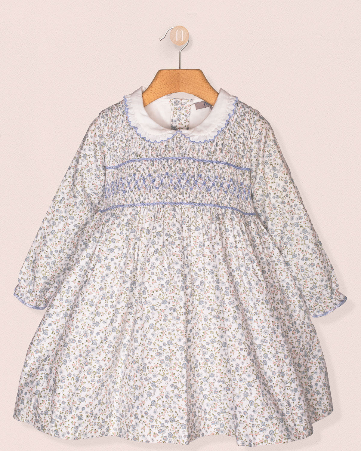 A front-facing view of the Fiorella Belina dress, featuring a delicate blue floral print, hand-smocked bodice, scalloped white collar, and long puffed sleeves with elastic cuffs. The dress is displayed on a wooden hanger against a soft pink wall.