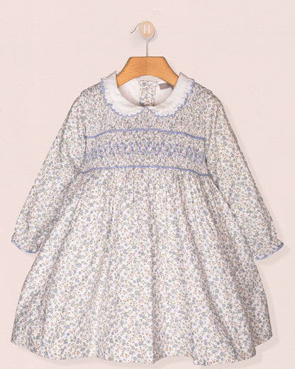 A front-facing view of the Fiorella Belina dress, featuring a delicate blue floral print, hand-smocked bodice, scalloped white collar, and long puffed sleeves with elastic cuffs. The dress is displayed on a wooden hanger against a soft pink wall.
