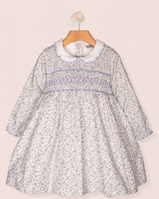 A front-facing view of the Fiorella Belina dress, featuring a delicate blue floral print, hand-smocked bodice, scalloped white collar, and long puffed sleeves with elastic cuffs. The dress is displayed on a wooden hanger against a soft pink wall.