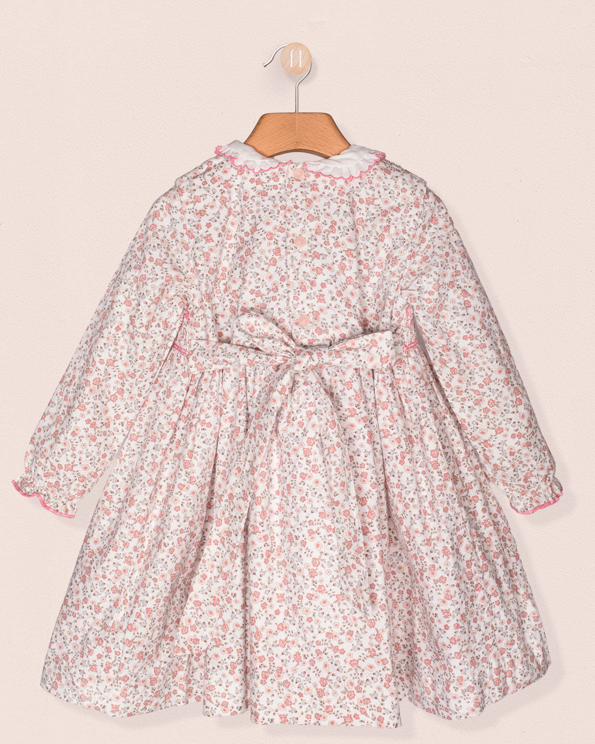 Back view of the same floral smocked dress, showing the self-tie bow at the waist and a gathered full skirt, displayed against a blush background.