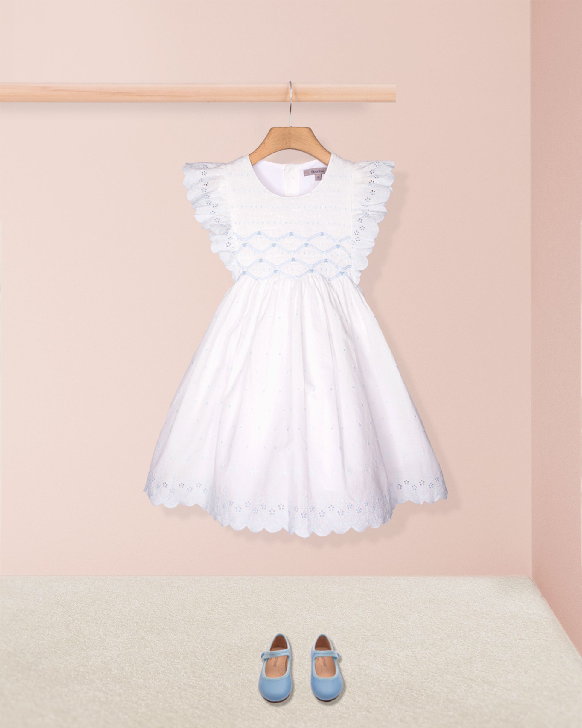White dress with ruffled sleeves on a hanger against a pink wall with blue shoes on the floor.