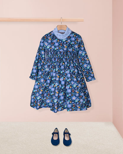 Blue floral dress on a hanger with matching shoes on the floor against a pink wall.