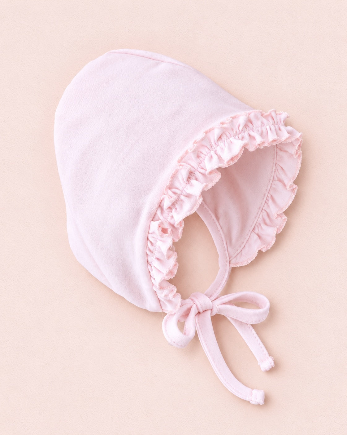 Pink baby's hat with a ruffled brim and tied around where the neck area would be if worn on a child, against a blush background