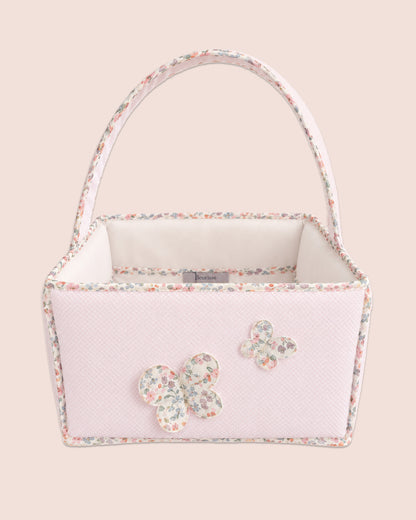 A light pink textured fabric basket bag with a floral print trim, a white interior lining, and two butterfly appliqués on the front, set against a solid pale pink background.