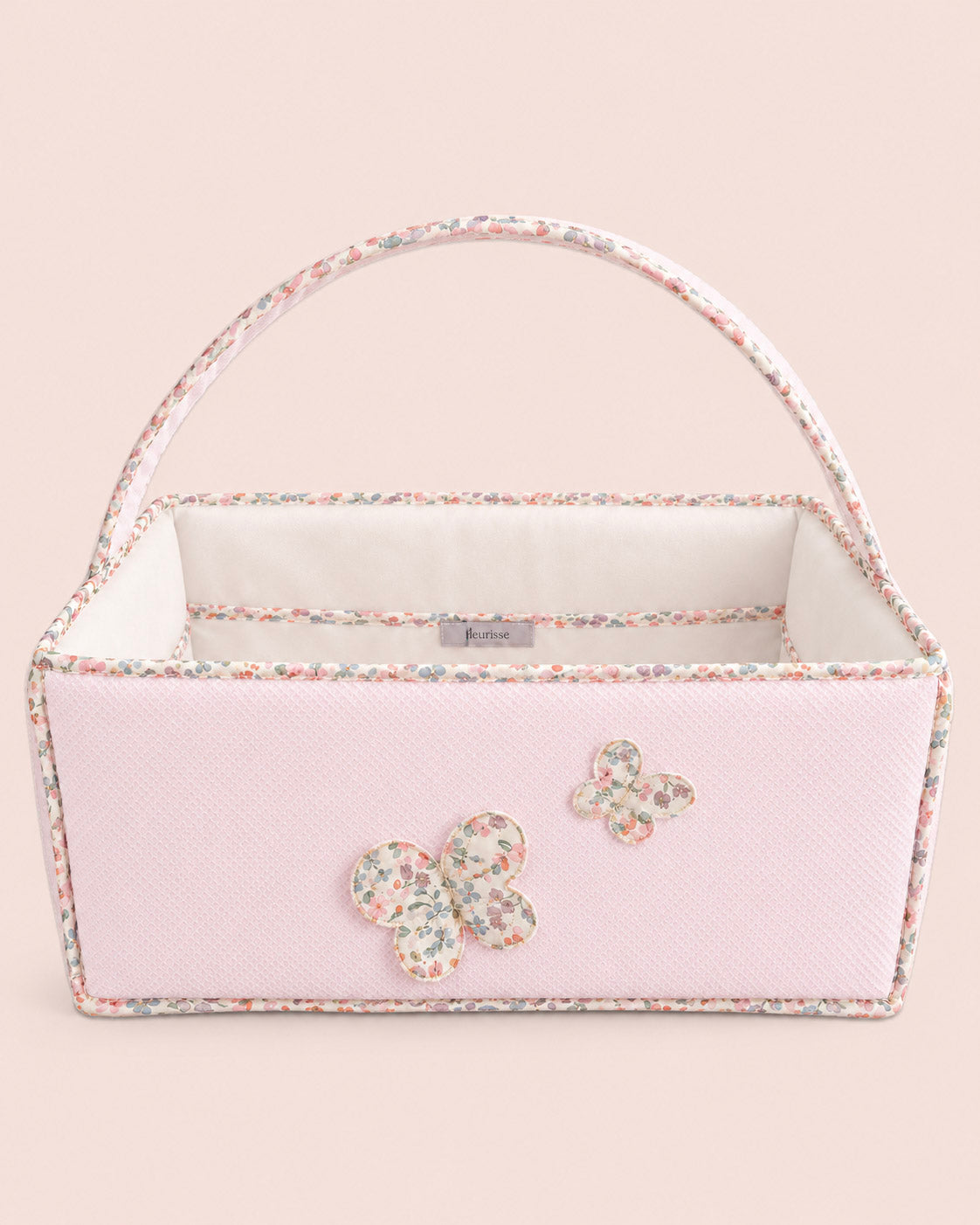 A light pink textured fabric storage basket with a floral trim, a white interior lining, and two butterfly appliqués on the front, set against a solid pale pink background.