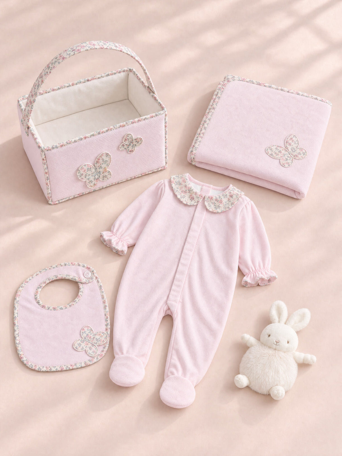 A coordinated baby gift set arranged on a soft blush background, including a light pink footed one-piece with a floral collar, a matching bib, a folded blanket, a small storage basket, and a white plush bunny toy.
