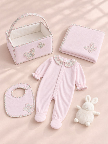 A coordinated baby gift set arranged on a soft blush background, including a light pink footed one-piece with a floral collar, a matching bib, a folded blanket, a small storage basket, and a white plush bunny toy.
