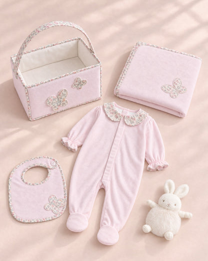 A coordinated baby gift set arranged on a soft blush background, including a light pink footed one-piece with a floral collar, a matching bib, a folded blanket, a small storage basket, and a white plush bunny toy.