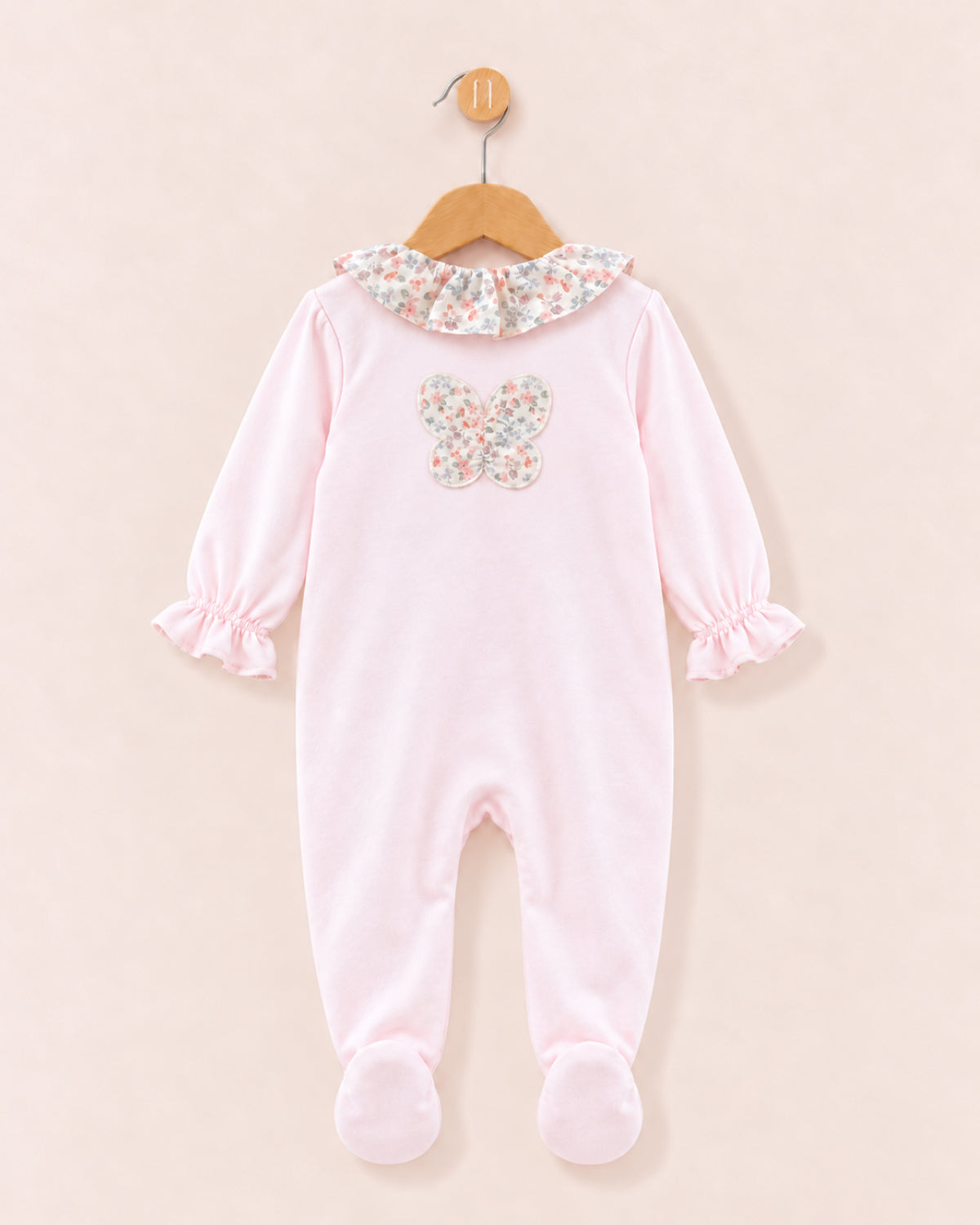 A baby footed sleeper in light pink, featuring a floral-patterned ruffled collar and a matching floral butterfly applique on the chest, hanging on a wooden hanger against a solid pale pink background.