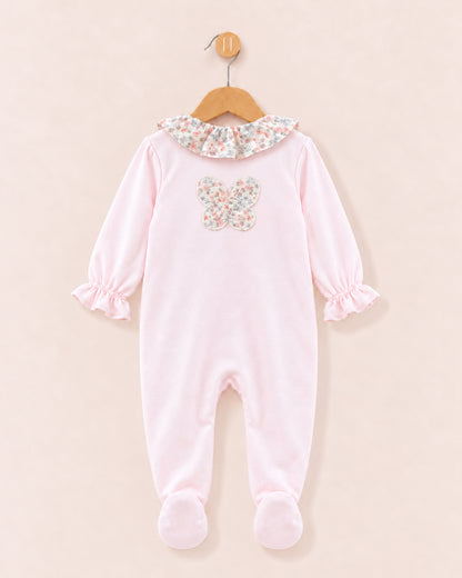 A baby footed sleeper in light pink, featuring a floral-patterned ruffled collar and a matching floral butterfly applique on the chest, hanging on a wooden hanger against a solid pale pink background.