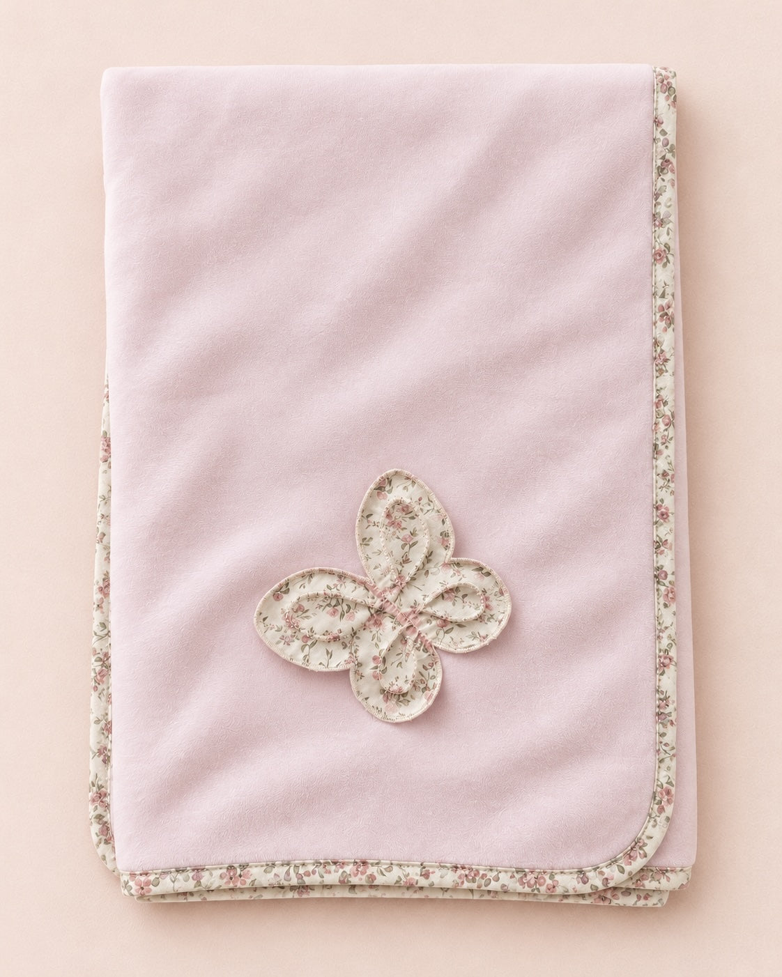 A folded light pink blanket with a floral patterned border and a matching embroidered butterfly applique in the center, displayed on a solid pale pink background.