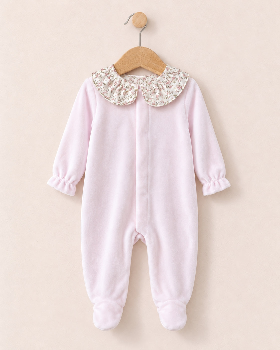 A baby girl's light pink footed sleeper with long sleeves and a decorative floral-patterned Peter Pan collar, hanging on a wooden hanger against a solid beige background.