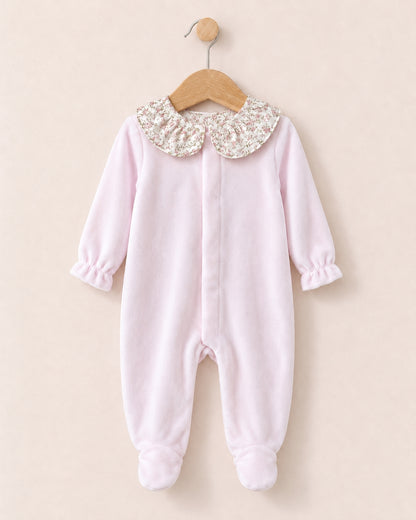 A baby girl's light pink footed sleeper with long sleeves and a decorative floral-patterned Peter Pan collar, hanging on a wooden hanger against a solid beige background.