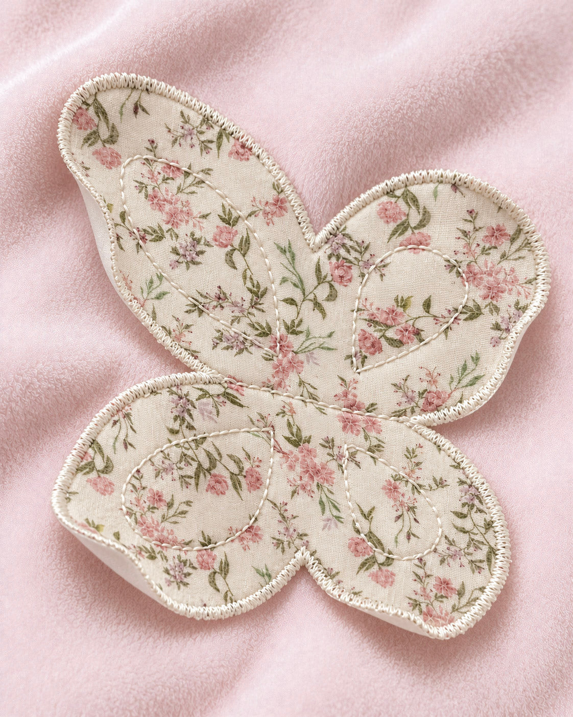 A butterfly-shaped applique patch with a pink and green floral pattern on a cream background, finished with a white embroidered border, laid flat on a soft pink fabric surface.