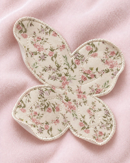 A butterfly-shaped applique patch with a pink and green floral pattern on a cream background, finished with a white embroidered border, laid flat on a soft pink fabric surface.