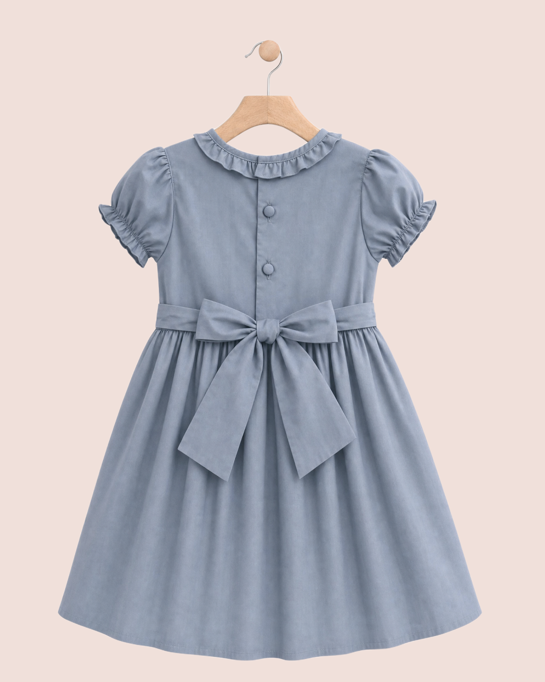 Back view of a blue-gray girl's dress with puff sleeves, a ruffled collar, and smocked details on the bodice, hanging on a wooden hanger against a solid pale pink background.