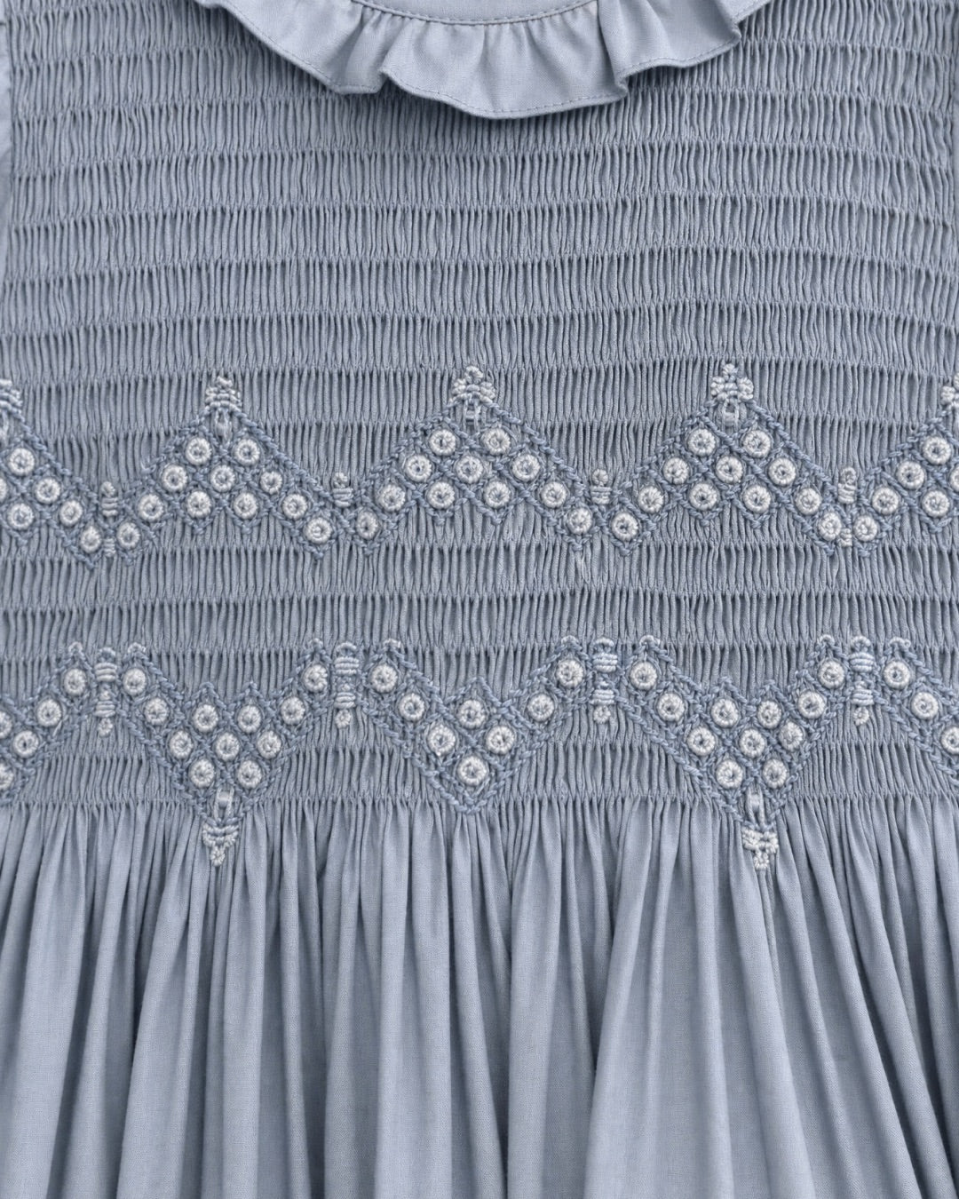 Close-up of a blue smocked dress featuring decorative embroidered diamond patterns with small white bead accents and a ruffled collar.