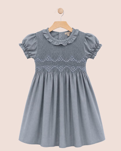 A blue-gray girl's dress with puff sleeves, a ruffled collar, and smocked details on the bodice, hanging on a wooden hanger against a solid pale pink background.