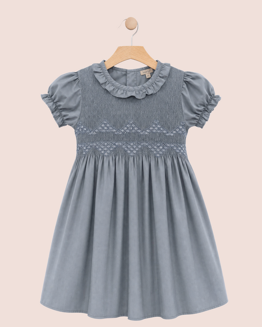 A blue-gray girl's dress with puff sleeves, a ruffled collar, and smocked details on the bodice, hanging on a wooden hanger against a solid pale pink background.