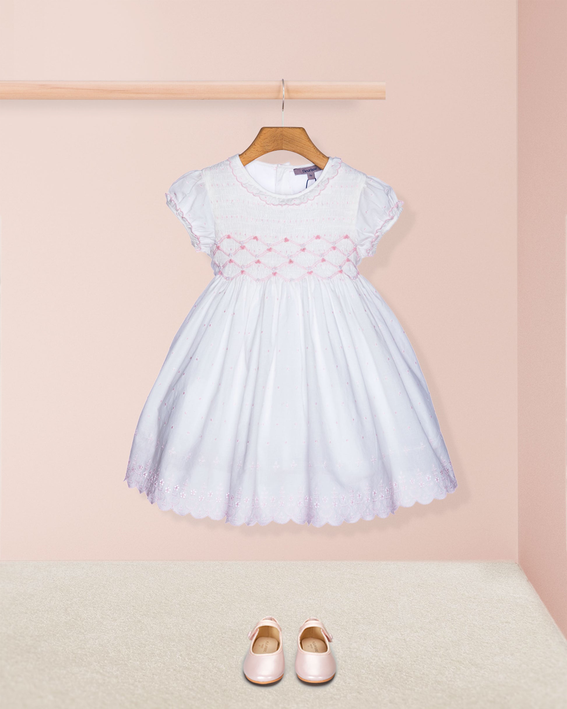 White dress with pink embroidery on a hanger against a pink wall, with pink shoes on the floor.