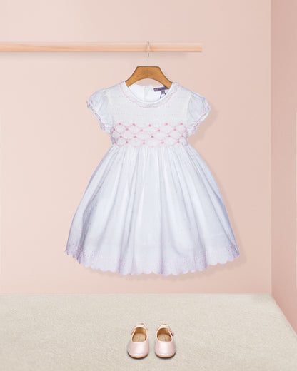 White dress with pink embroidery on a hanger against a pink wall, with pink shoes on the floor.