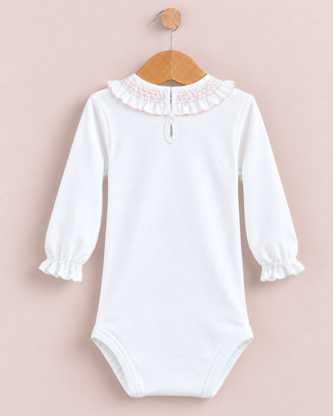A white long-sleeve baby bodysuit with a ruffled lace collar, hanging on a wooden hanger against a soft pink background.