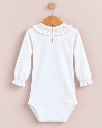 A white long-sleeve baby bodysuit with a ruffled lace collar, hanging on a wooden hanger against a soft pink background.