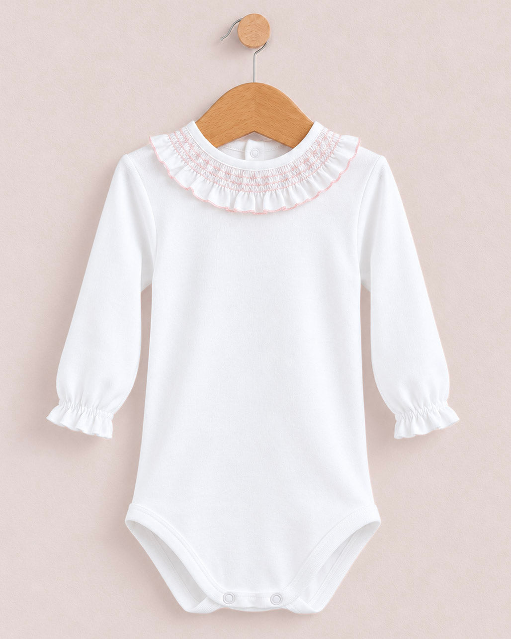 A white long-sleeve baby bodysuit with a ruffled lace collar and elastic cuffs, hanging on a wooden hanger against a soft pink background.