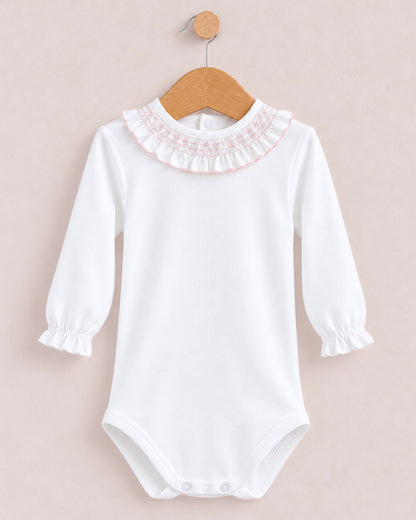 A white long-sleeve baby bodysuit with a ruffled lace collar and elastic cuffs, hanging on a wooden hanger against a soft pink background.