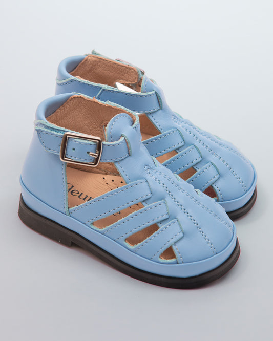 Pair of baby boy light blue leather sandals with open-weave front, ankle strap, and velcro closure with silver buckle, displayed on a neutral gray background.