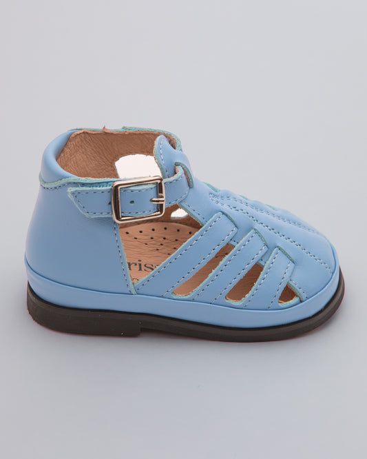 Side view of a light blue leather baby boy sandal with rubber sole, open side cutouts, and velcro ankle strap with decorative buckle, placed on a gray surface.