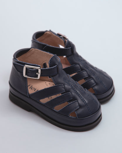 Pair of navy blue leather baby boy sandals with open-weave design, velcro ankle strap, and silver buckle, displayed on a soft grey background.